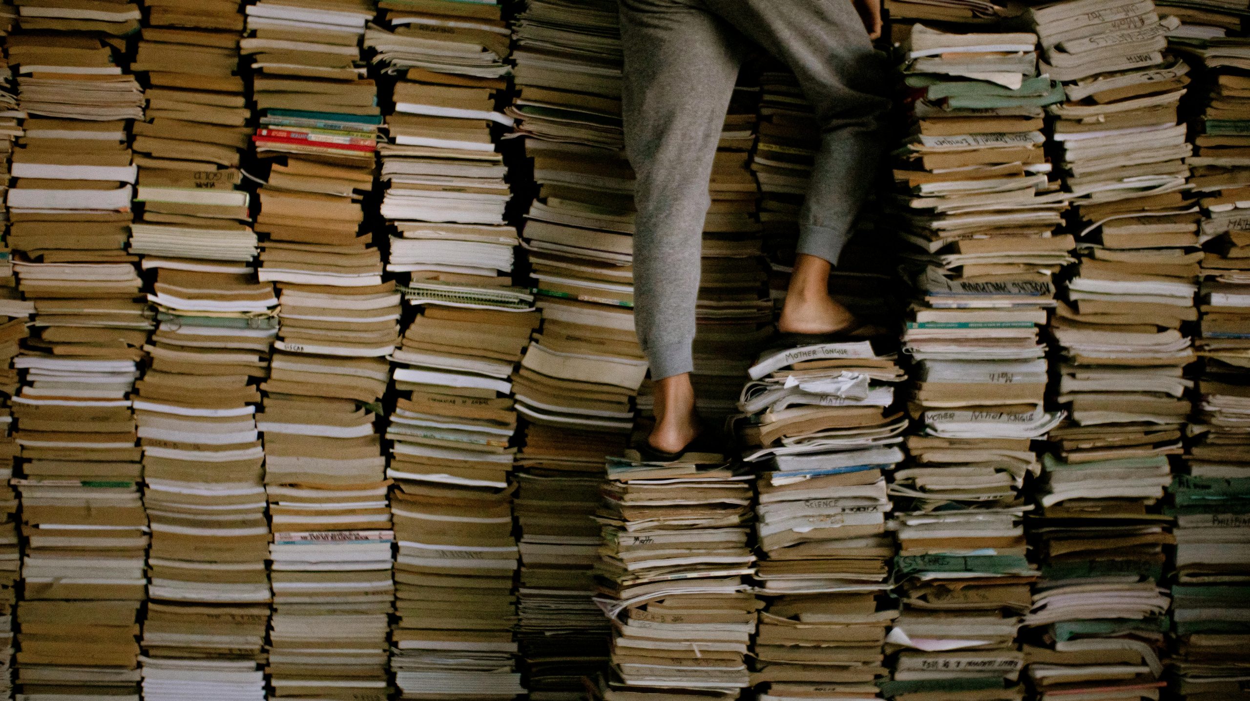 A person in gray sweatpants climbs on large stacks of old books indoors.